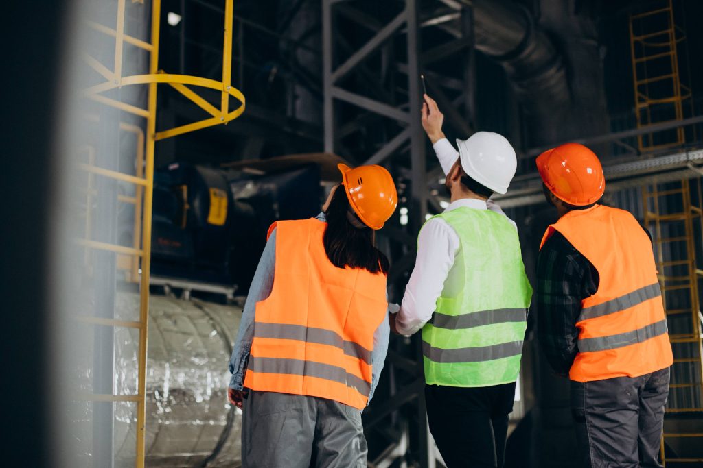 Un grupo de tres profesionales de la industria minera, equipados con cascos de seguridad (naranjas y blancos) y chalecos reflectantes (naranjas y verde), colaboran e inspeccionan juntos maquinaria y estructuras metálicas en una instalación industrial. El supervisor central señala hacia una estructura elevada, ilustrando la comunicación y el aprendizaje conjunto como parte del proceso de transformación cultural en minería.
